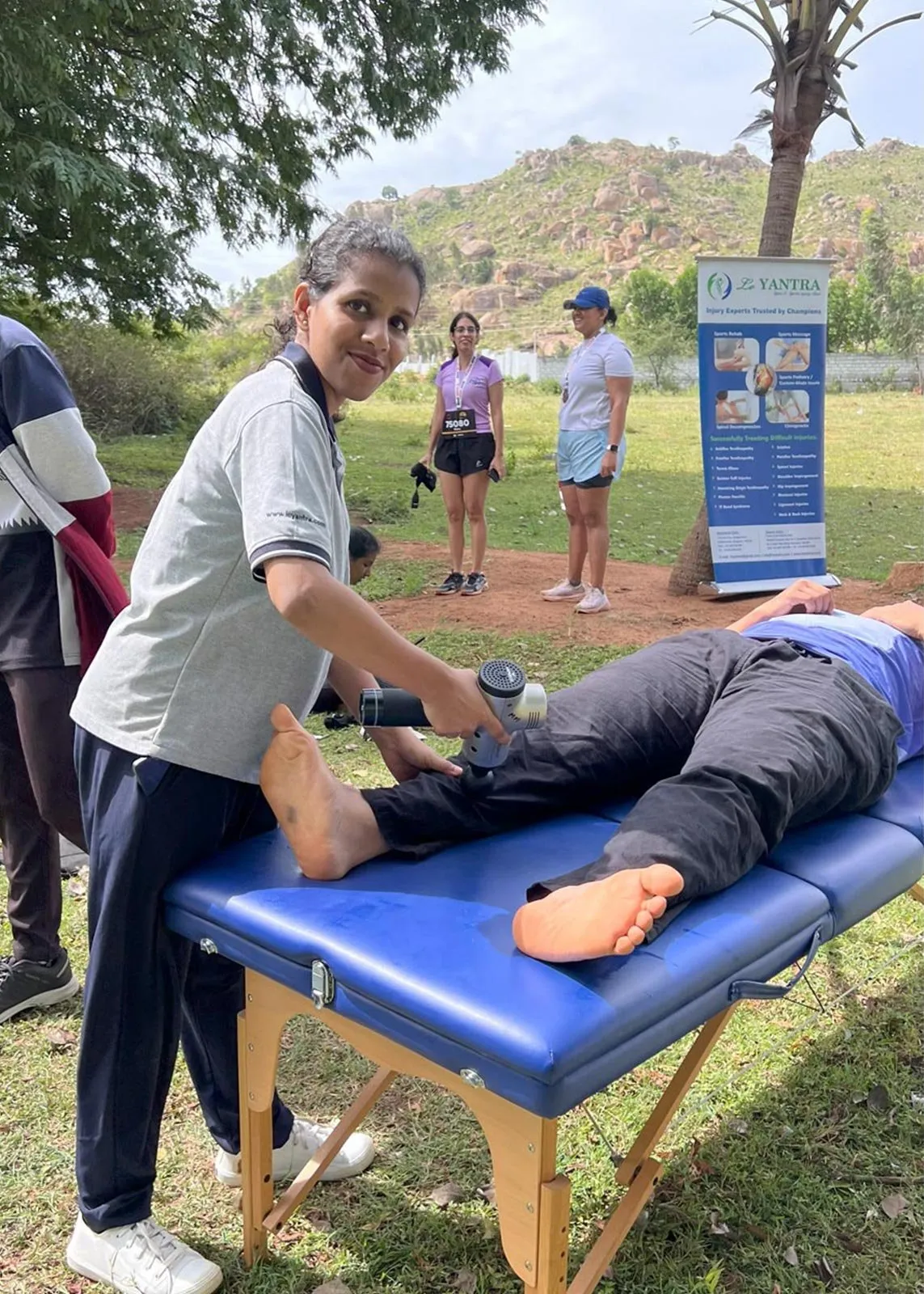A Le Yantra physiotherapist in a branded polo shirt using a percussion massage device on the lower leg of a person lying face down on a blue treatment couch, at an outdoor event with a Le Yantra banner and a rocky hill behind.
