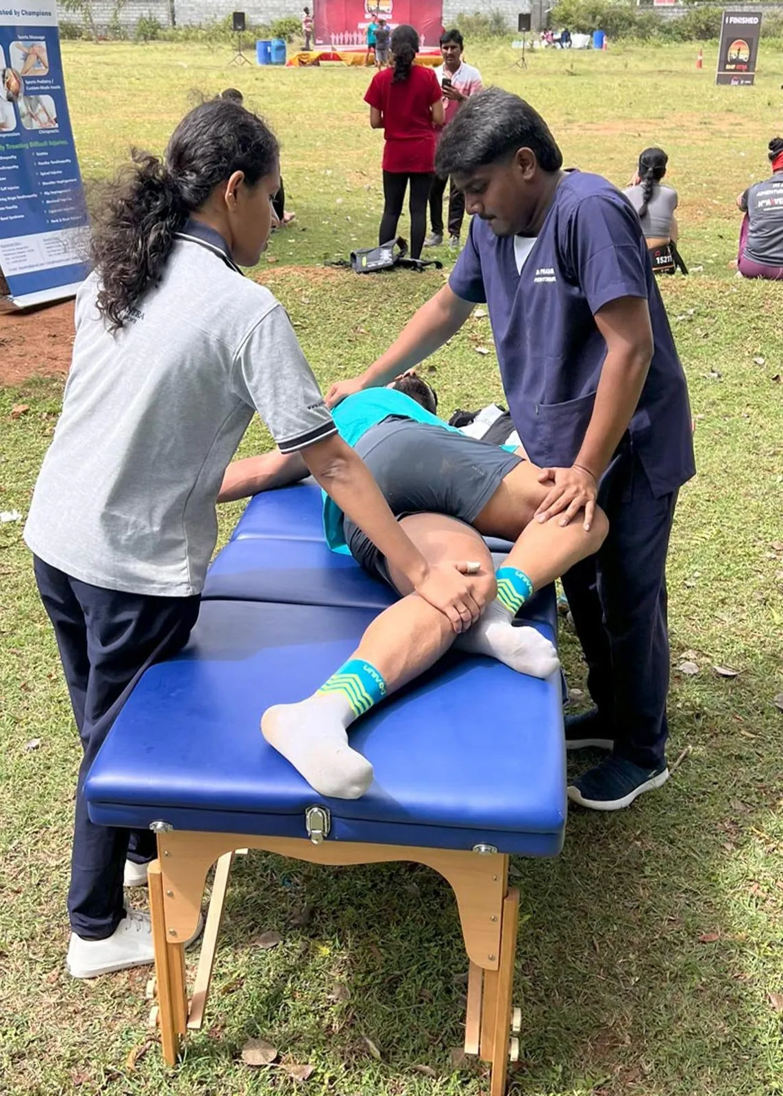 Two Le Yantra physiotherapists working together on a runner lying face down on a treatment couch at an event finish area, one supporting the shoulder and one flexing the knee.