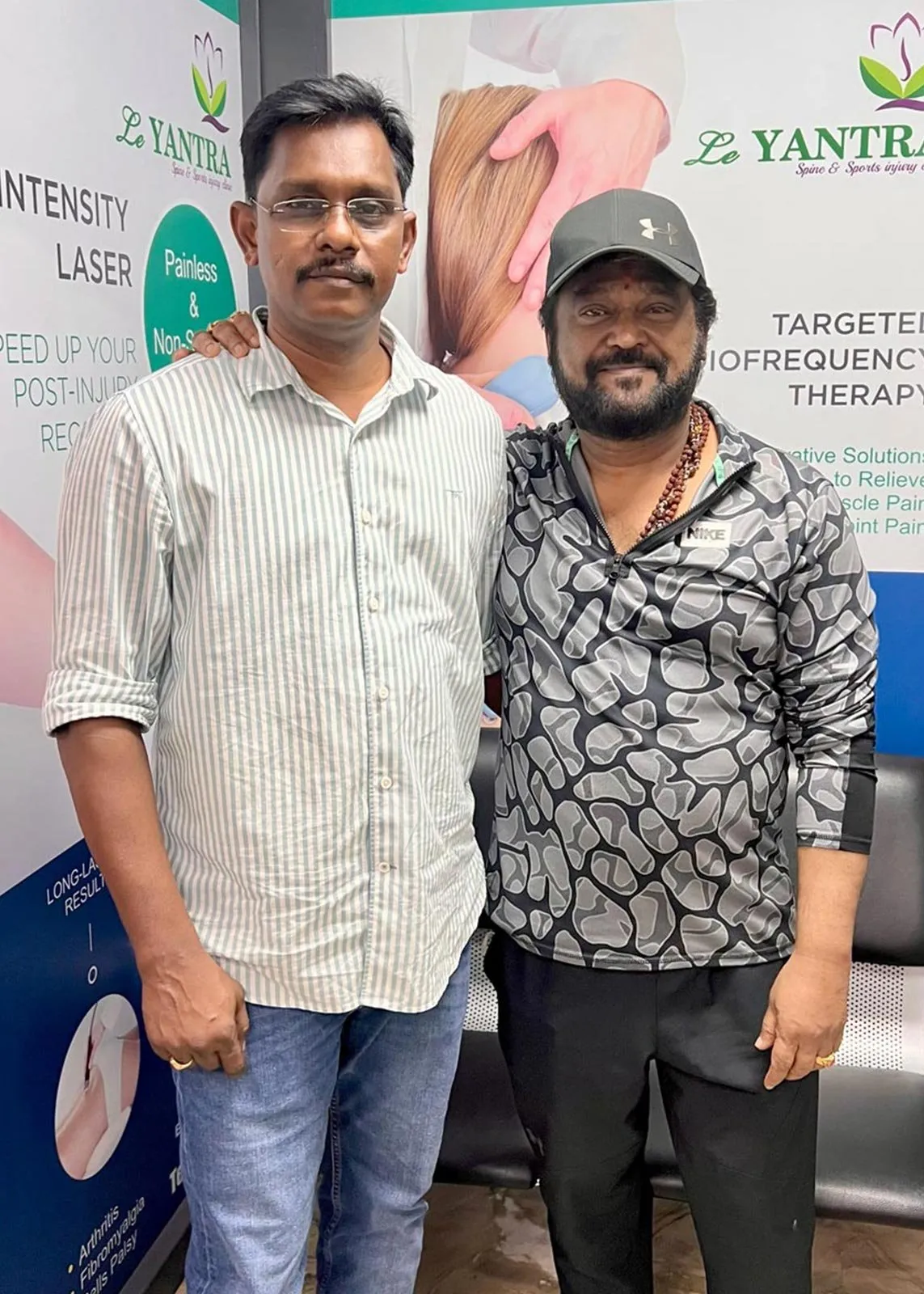 Two men standing side by side in front of Le Yantra banners for high-intensity laser and radiofrequency therapy inside the clinic.