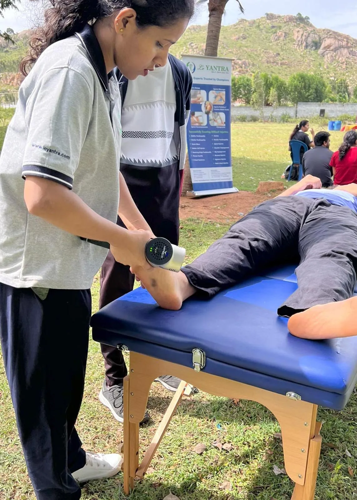 A Le Yantra physiotherapist using a percussion massage device on the calf of a person lying face down on a treatment couch, with a Le Yantra roll-up banner behind and an open field beyond.