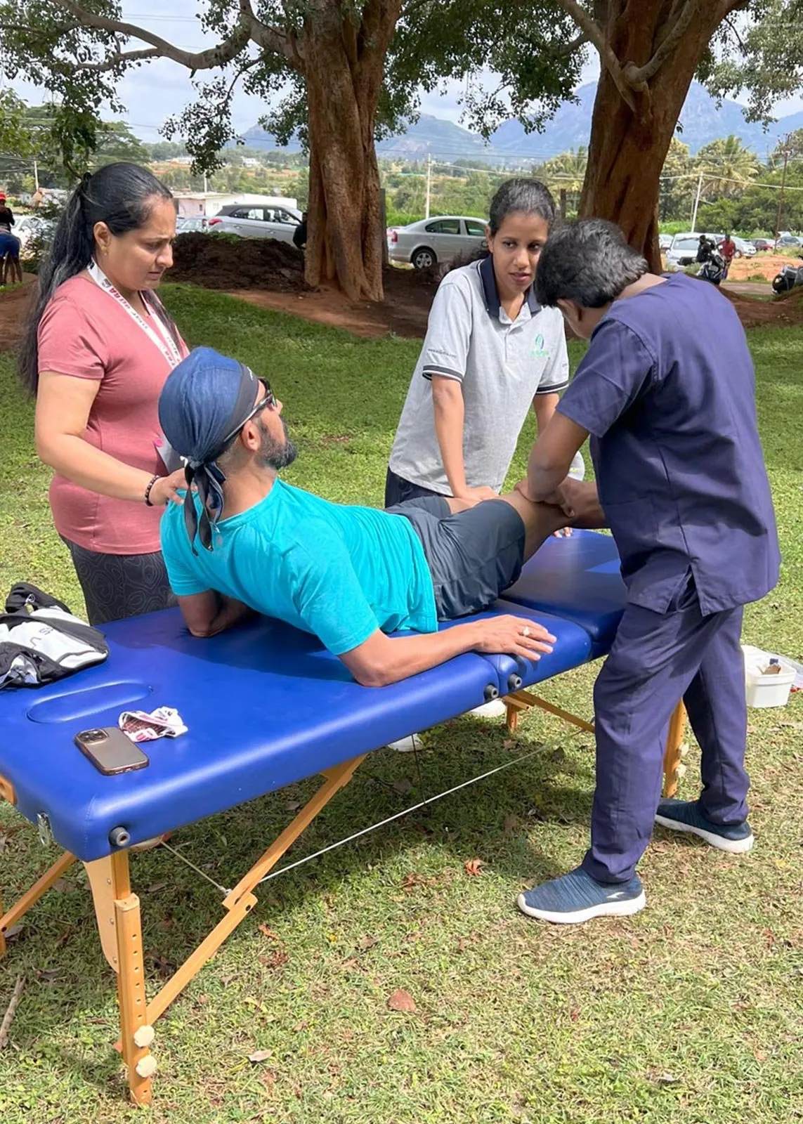 Three clinicians attending to a person lying face down on a treatment couch under trees at an outdoor event, one holding the knee and one steadying the shoulder.