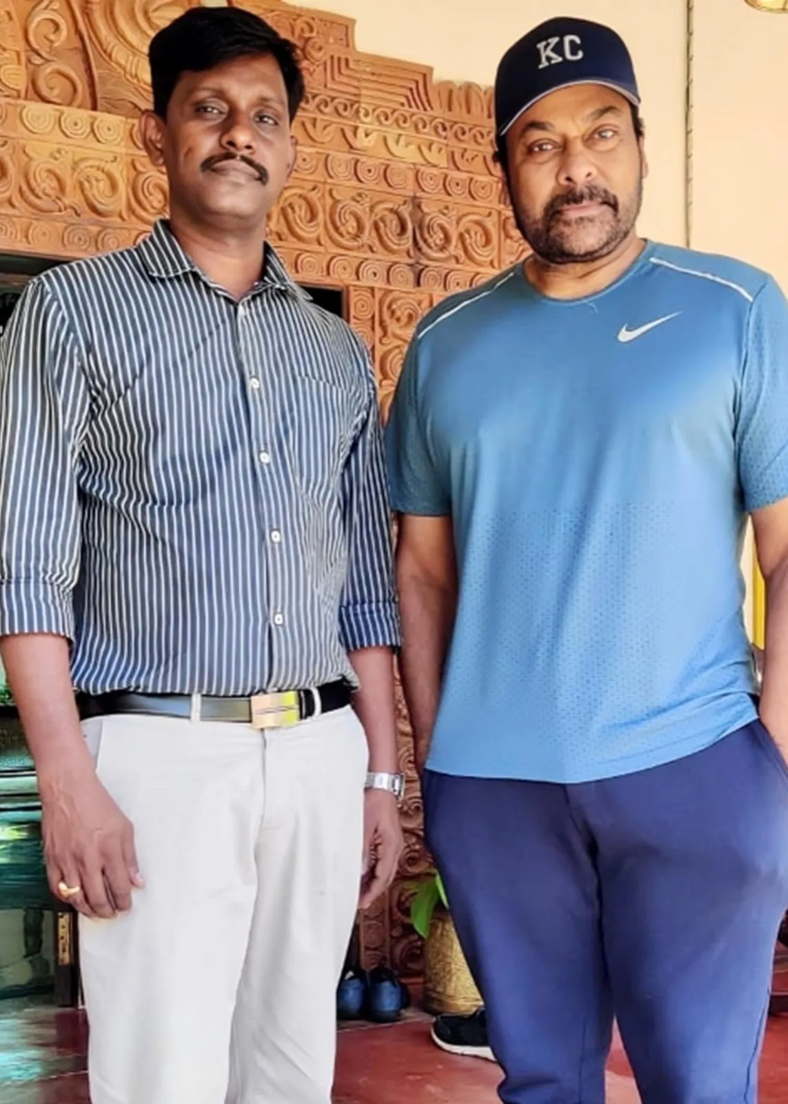 Two men standing side by side in a doorway with a carved wooden panel behind them.
