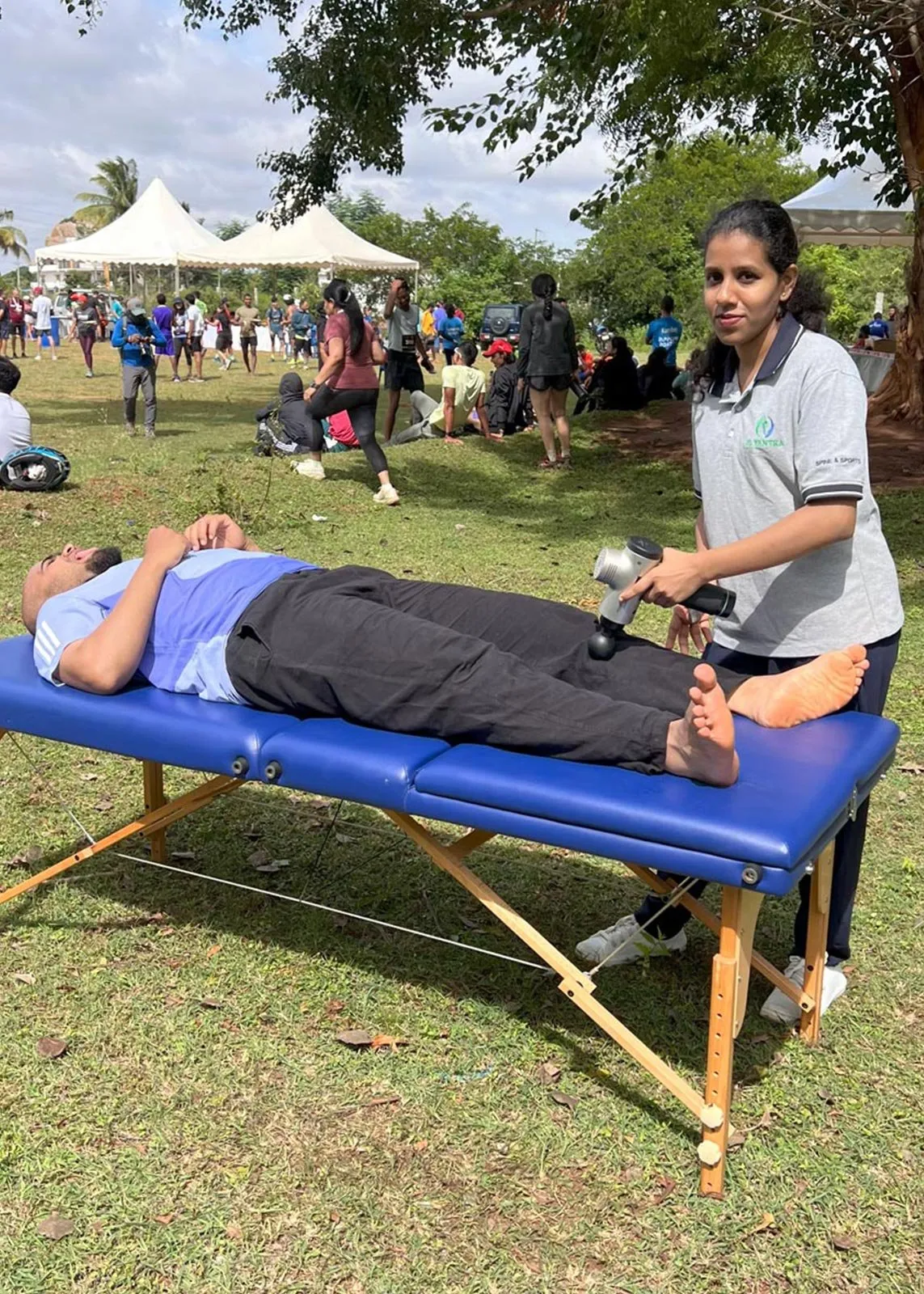 A Le Yantra physiotherapist using a percussion massage device on the foot of a person lying on their back on a treatment couch, with event tents and runners in the background.